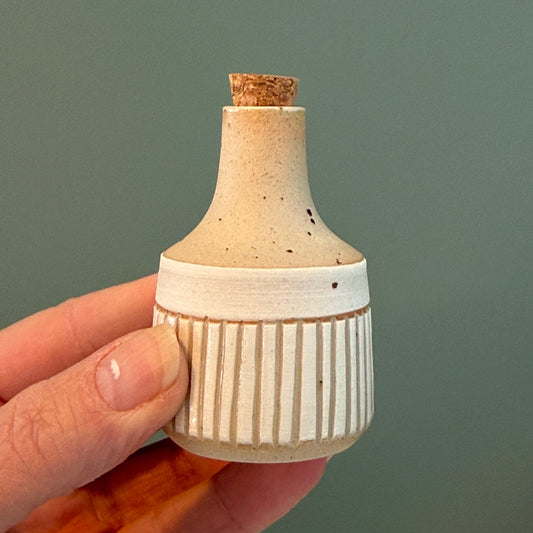 Hand holding a small ceramic bottle with a cork against a plain background