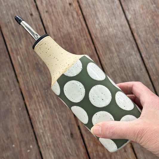 Hand holding an oil bottle with a green and white polka dot design on a wooden surface