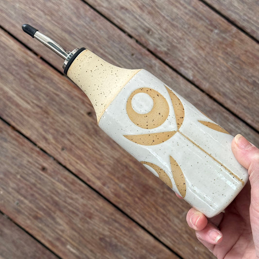 Decorative oil bottle with a beige and brown flower design held by a hand on a wooden surface