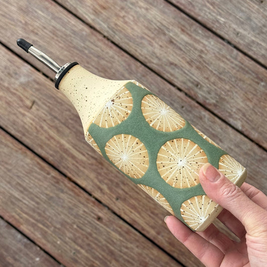 Hand holding a bottle with a decorative radial glaze pattern in white on green glaze, on a wooden surface